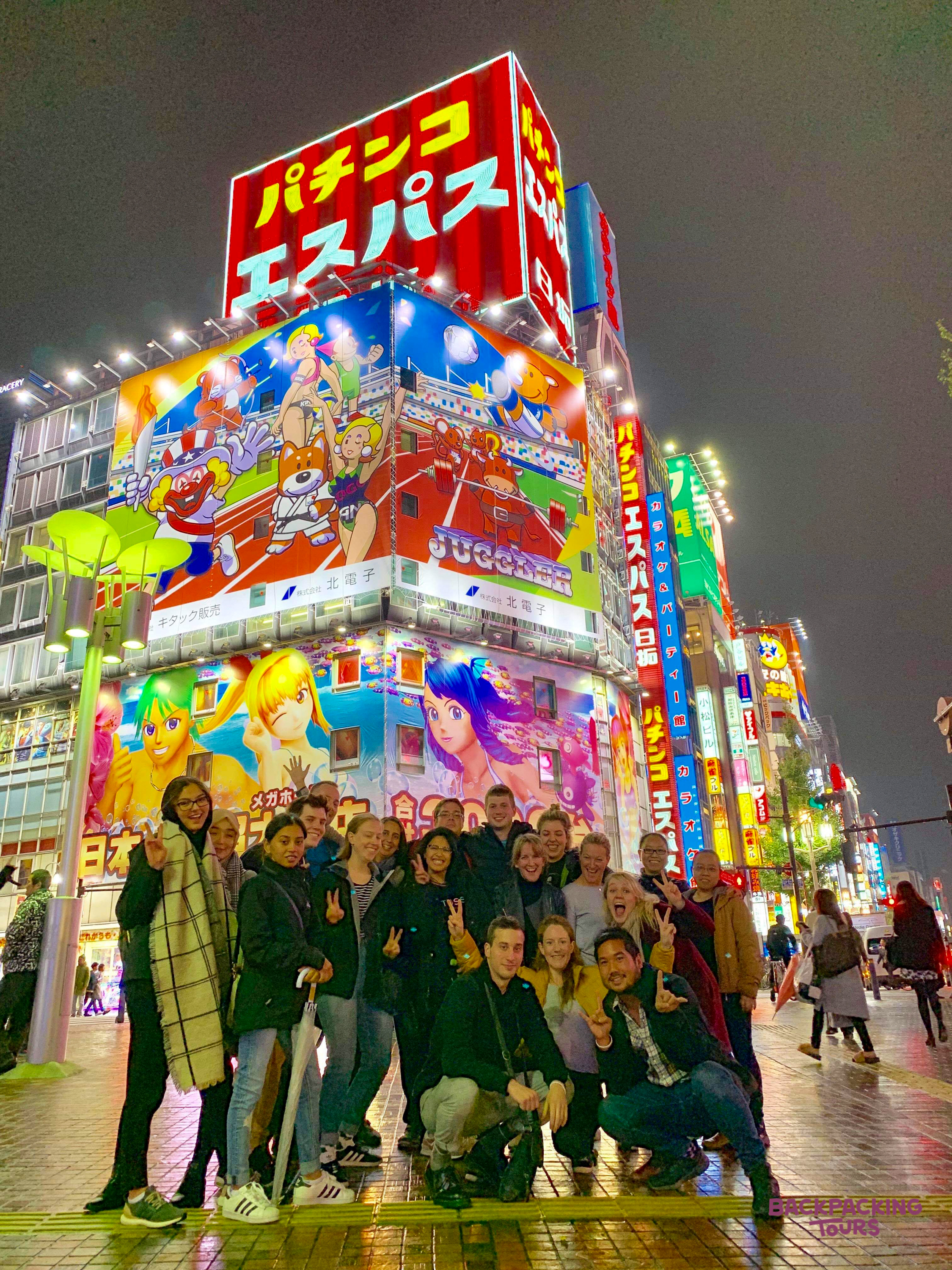 Happy backpackers pose on a wet street in Tokyo, surrounded by vibrant neon and anime signs at night.