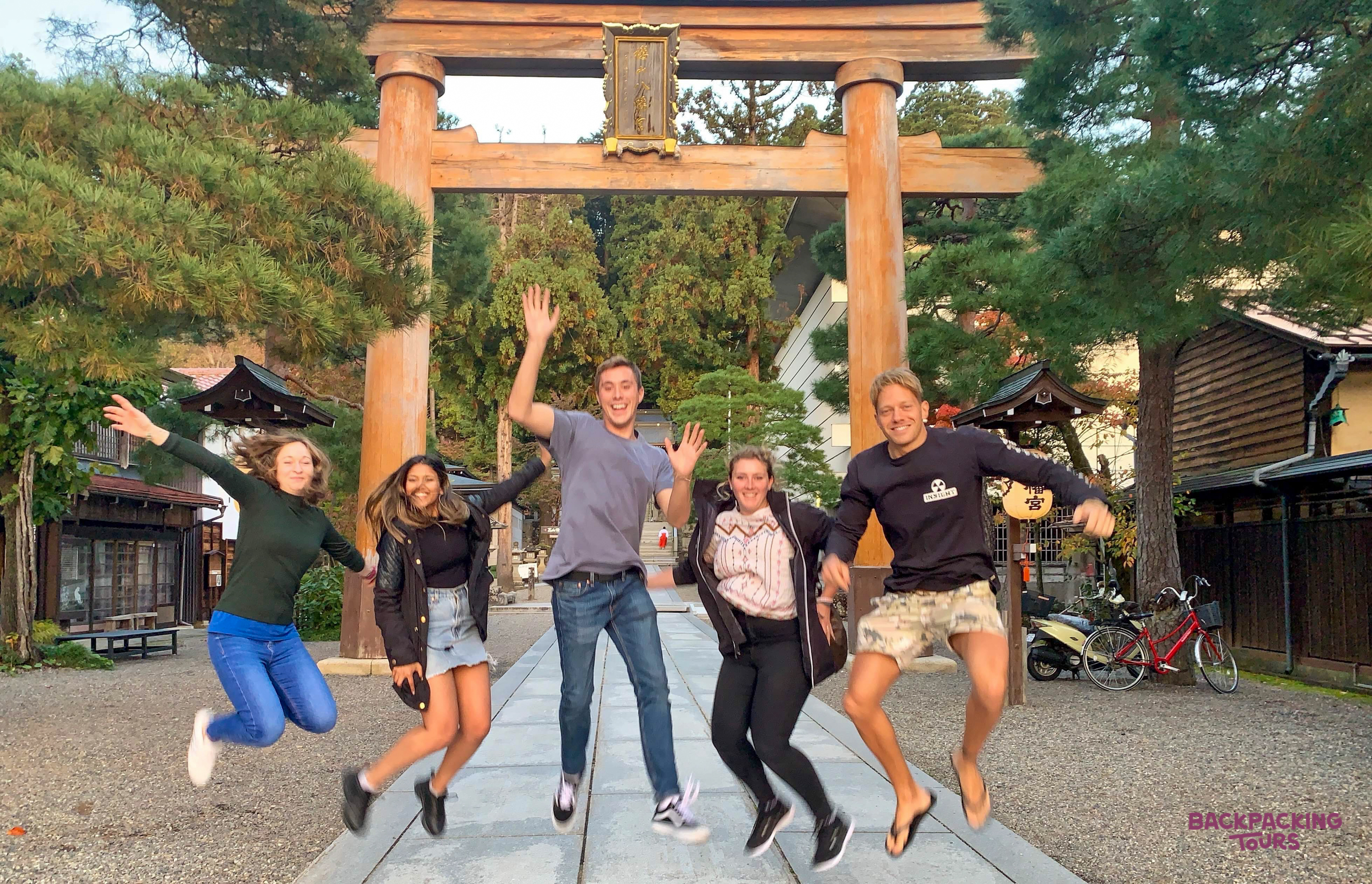 Five happy backpackers jump for joy in front of a traditional wooden torii gate at a shrine in Tokyo.