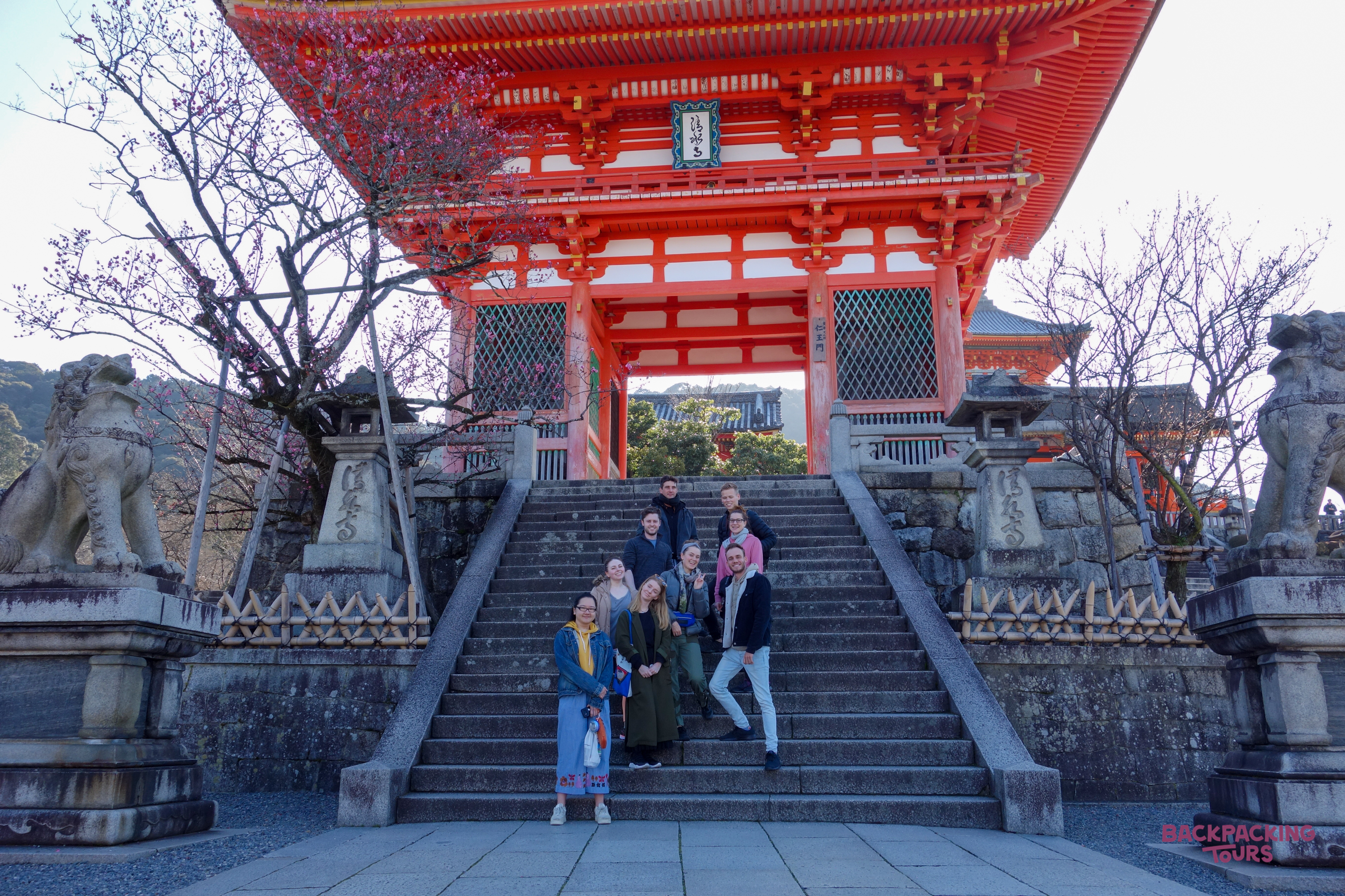 Backpackers pose on stone steps before the vibrant red Nio-mon gate of Kiyomizu-dera Temple in Kyoto.