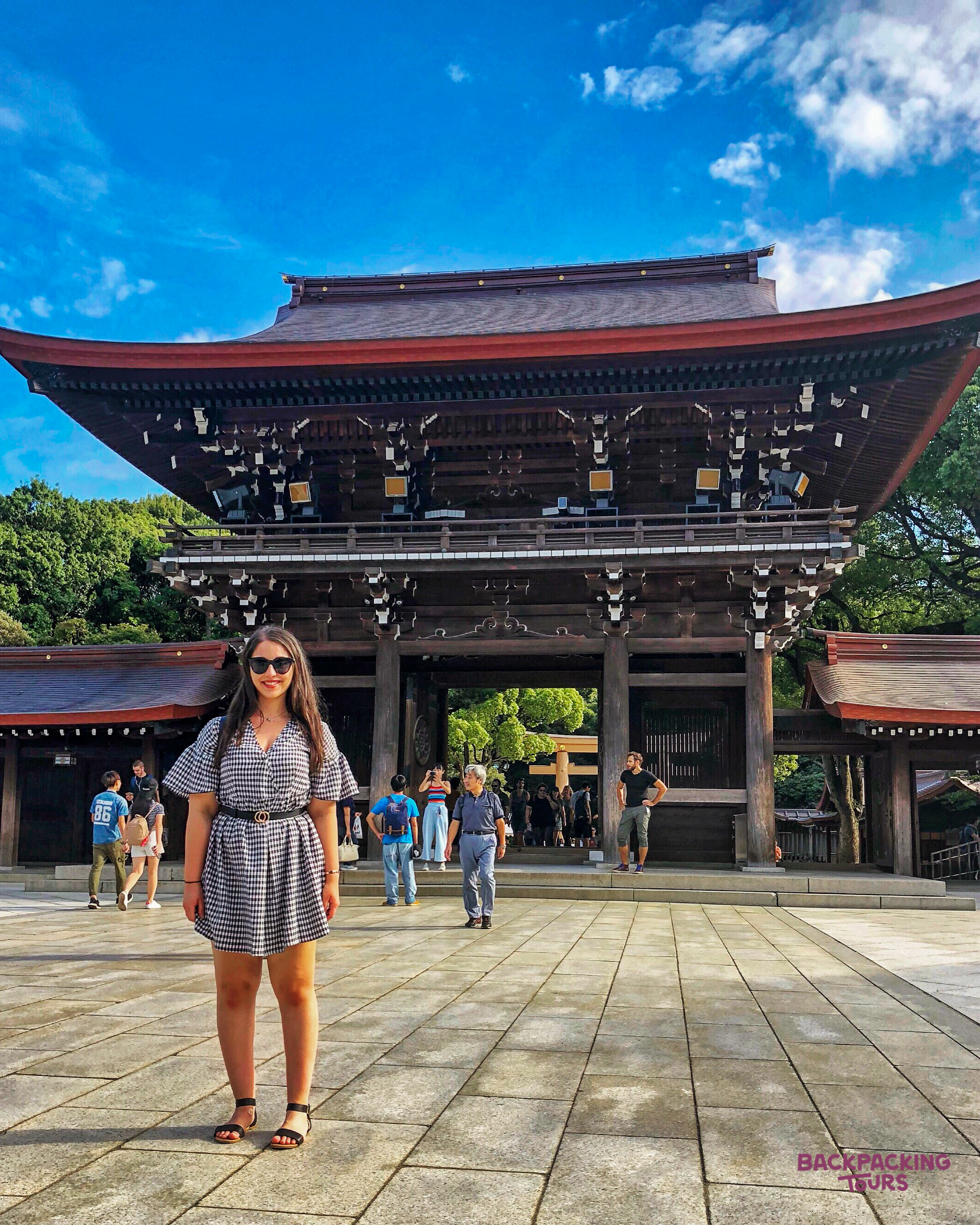 A smiling woman stands before a grand traditional Japanese temple gate in Kyoto under a blue sky.