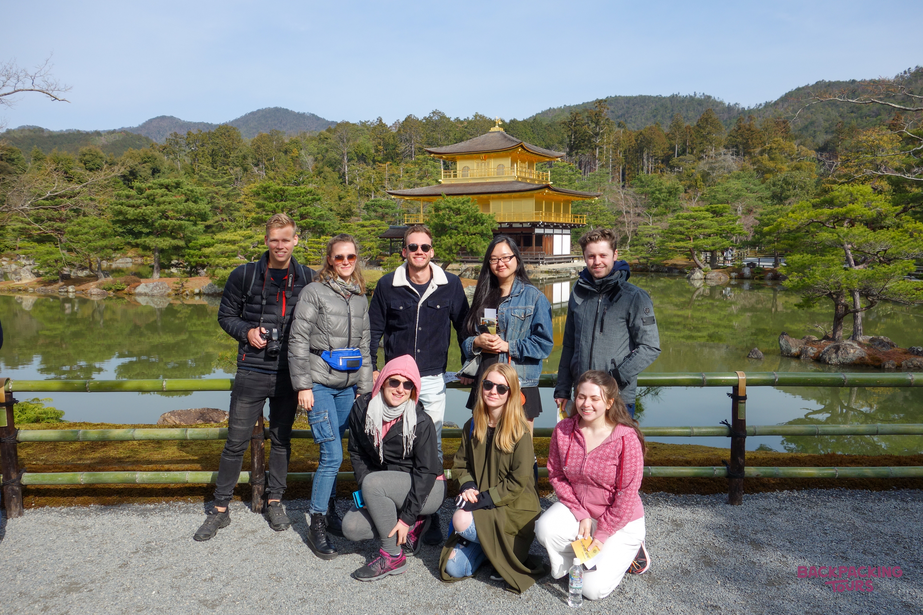 Smiling backpackers pose before Kyoto's iconic Golden Pavilion, reflected beautifully in the tranquil lake.