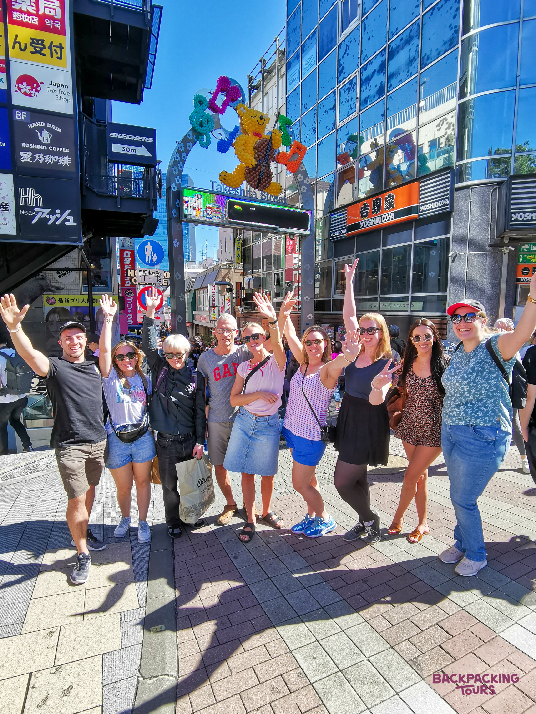 A group of happy backpackers waving on a bustling street in Harajuku, Tokyo, under a colorful archway.
