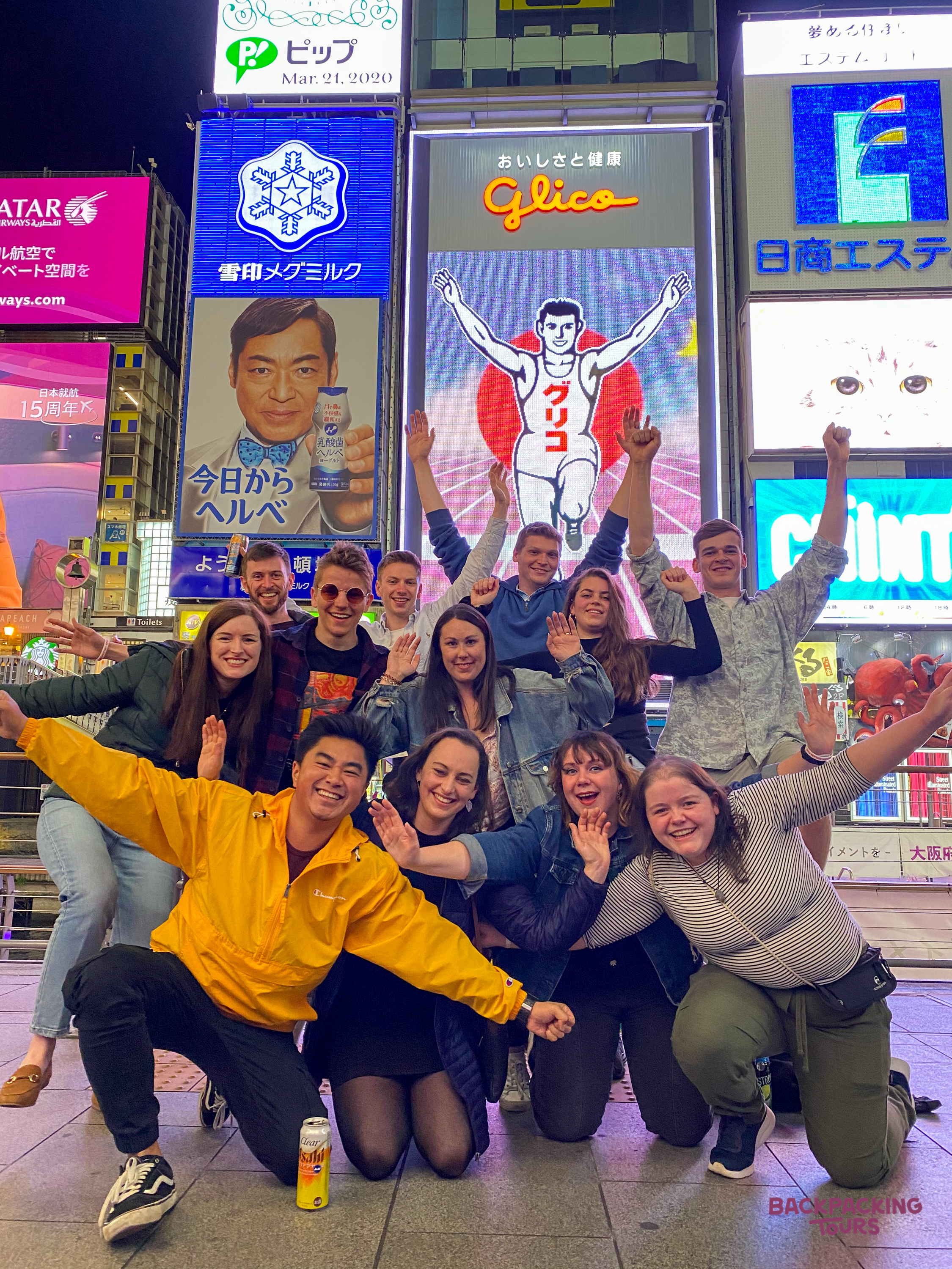 A joyful group of backpackers pose enthusiastically in front of the vibrant Glico sign in Dotonbori, Osaka.