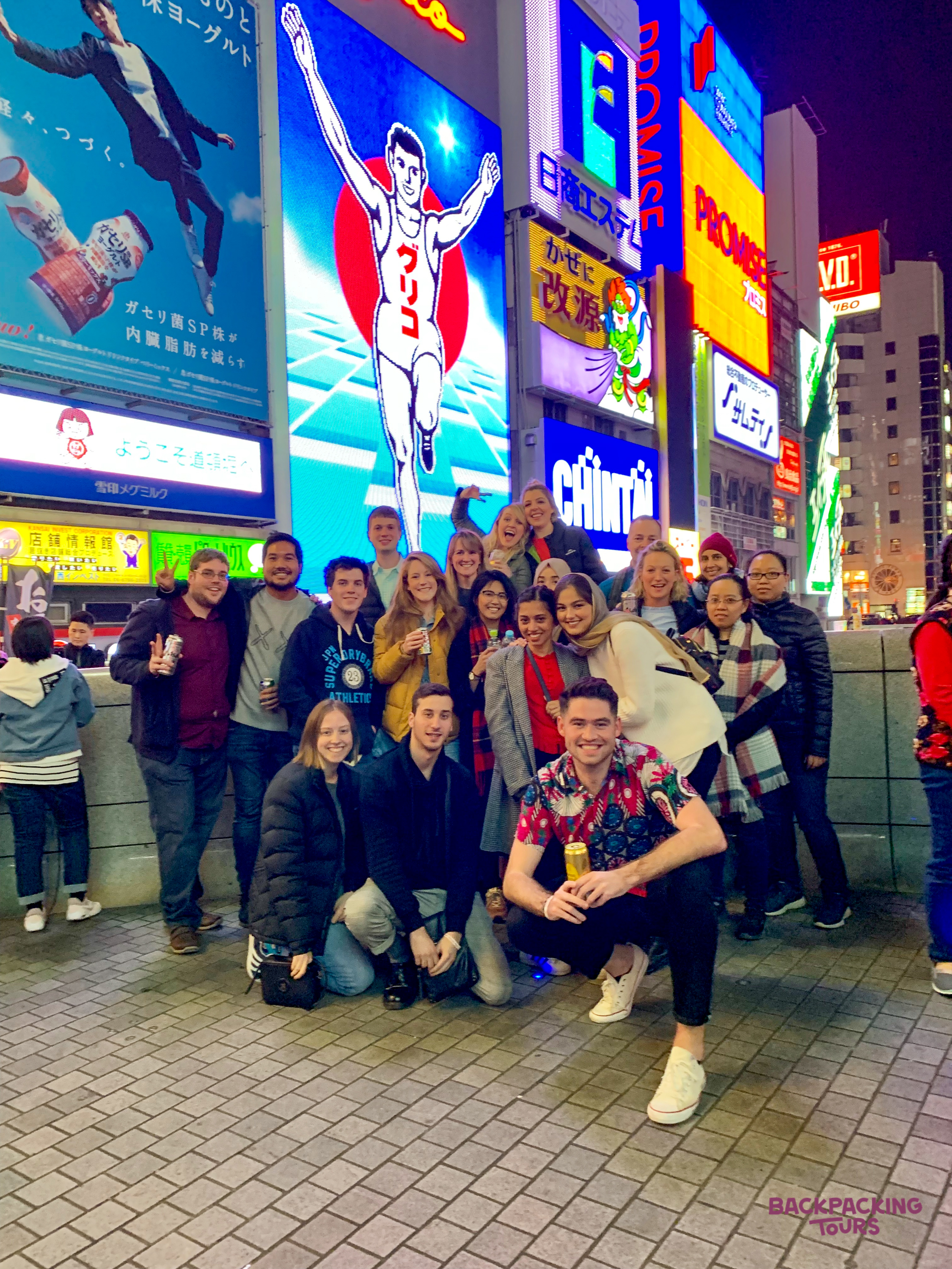 A happy group of backpackers poses at night in front of the iconic Glico Man billboard in Dotonbori, Osaka.