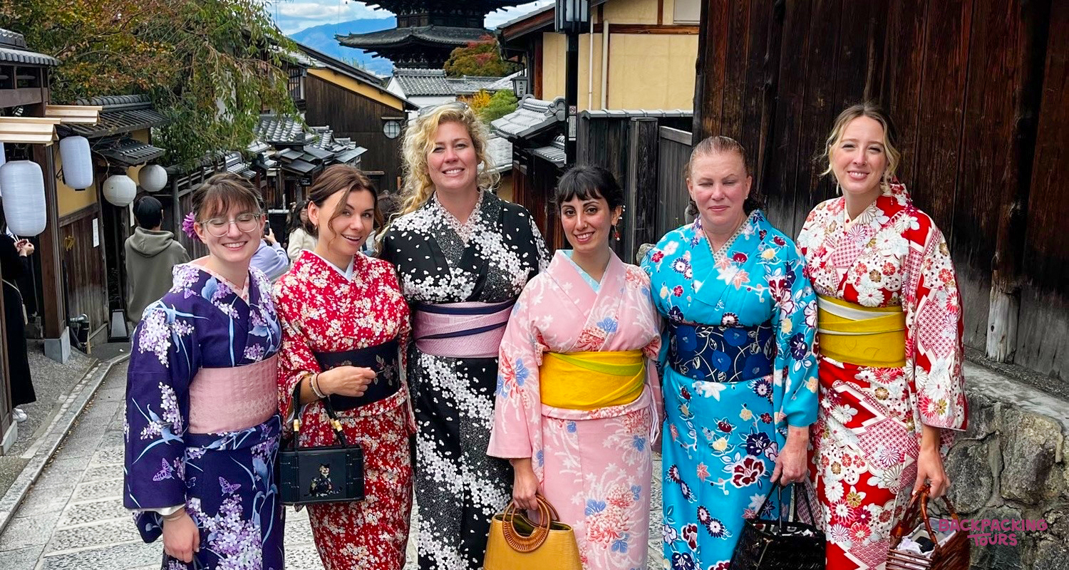 Six smiling women in colorful kimonos pose on a charming traditional street in Kyoto.