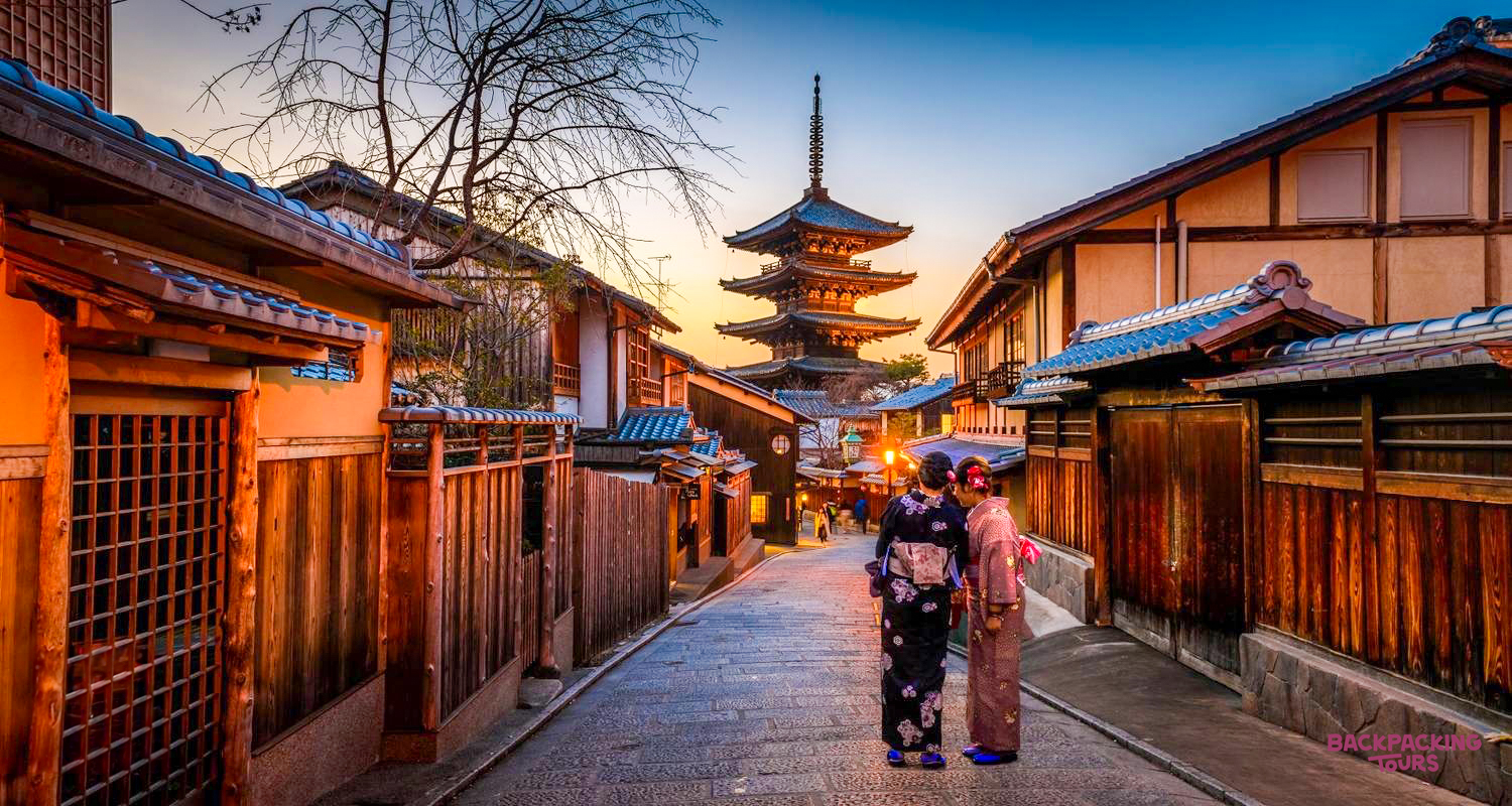 Two people in kimonos stroll along a traditional Kyoto street at sunset, a pagoda in the distance.