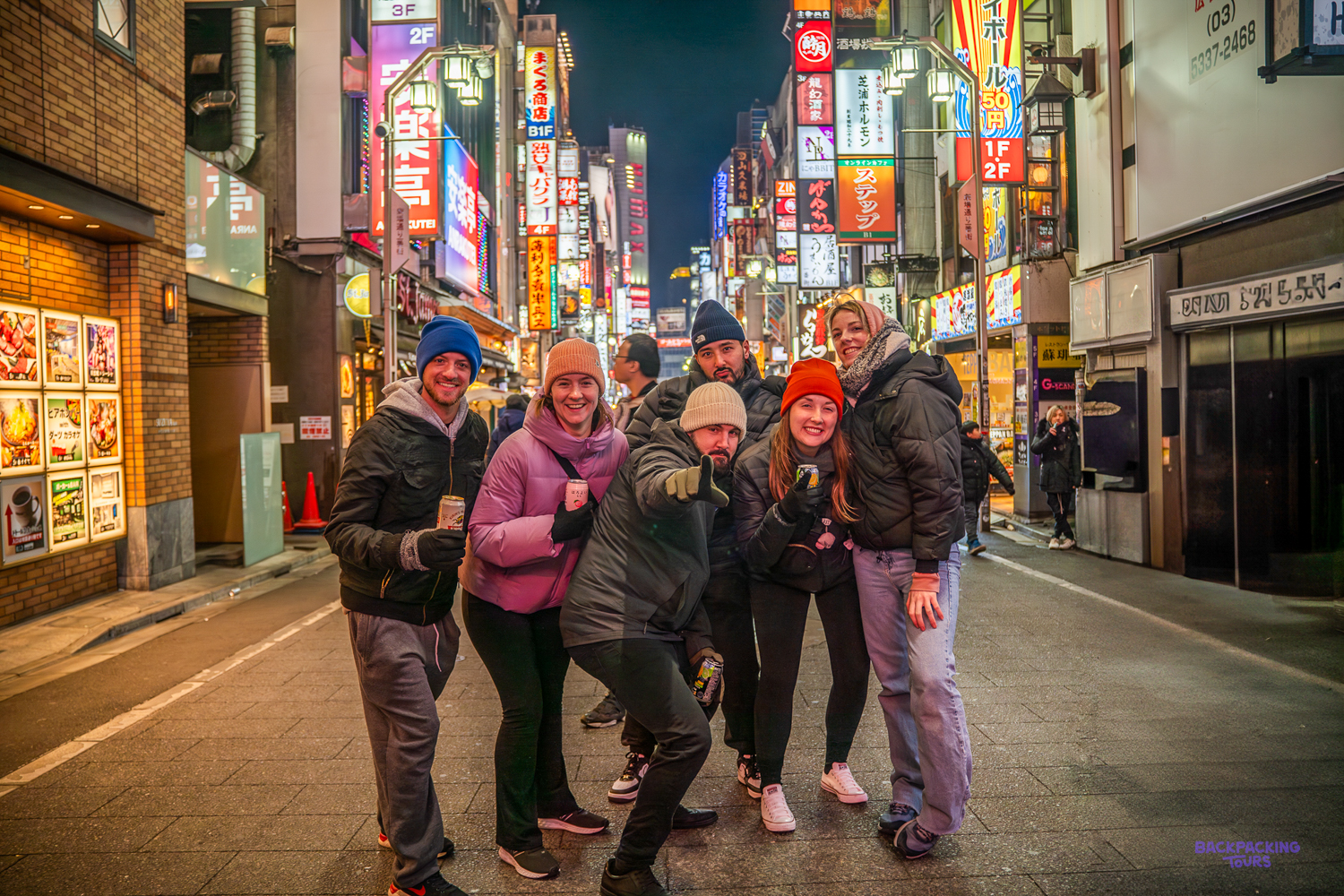 Group of backpackers smiling on a neon-lit Tokyo street on their winter welcome night.
