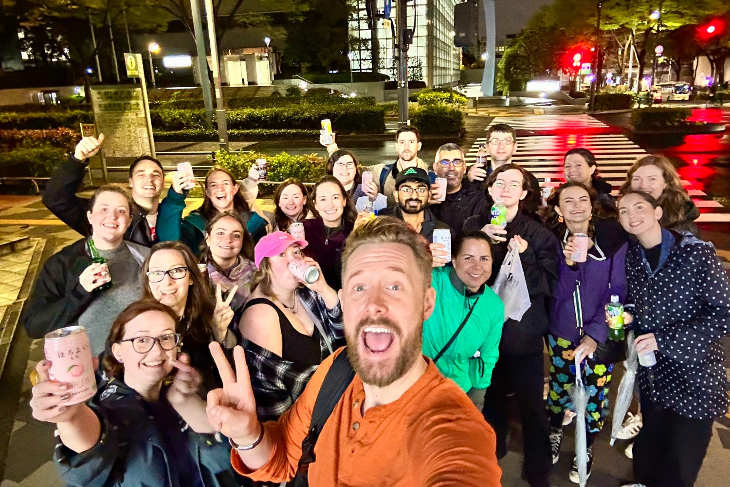 A joyful group of friends taking a selfie on a wet city street at night, holding drinks.