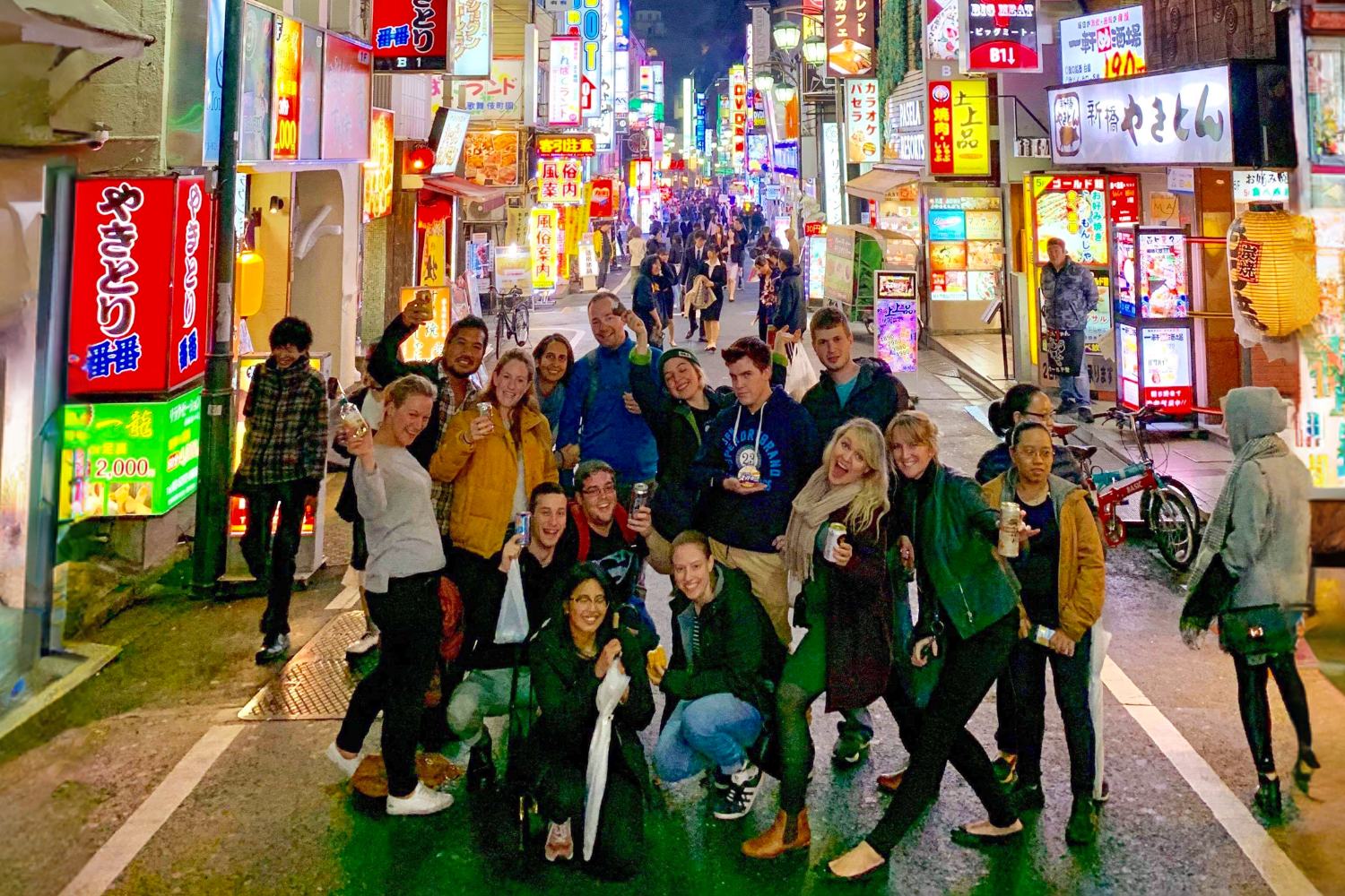 Smiling backpackers pose on a vibrant, neon-lit street in a lively Japanese city at night.