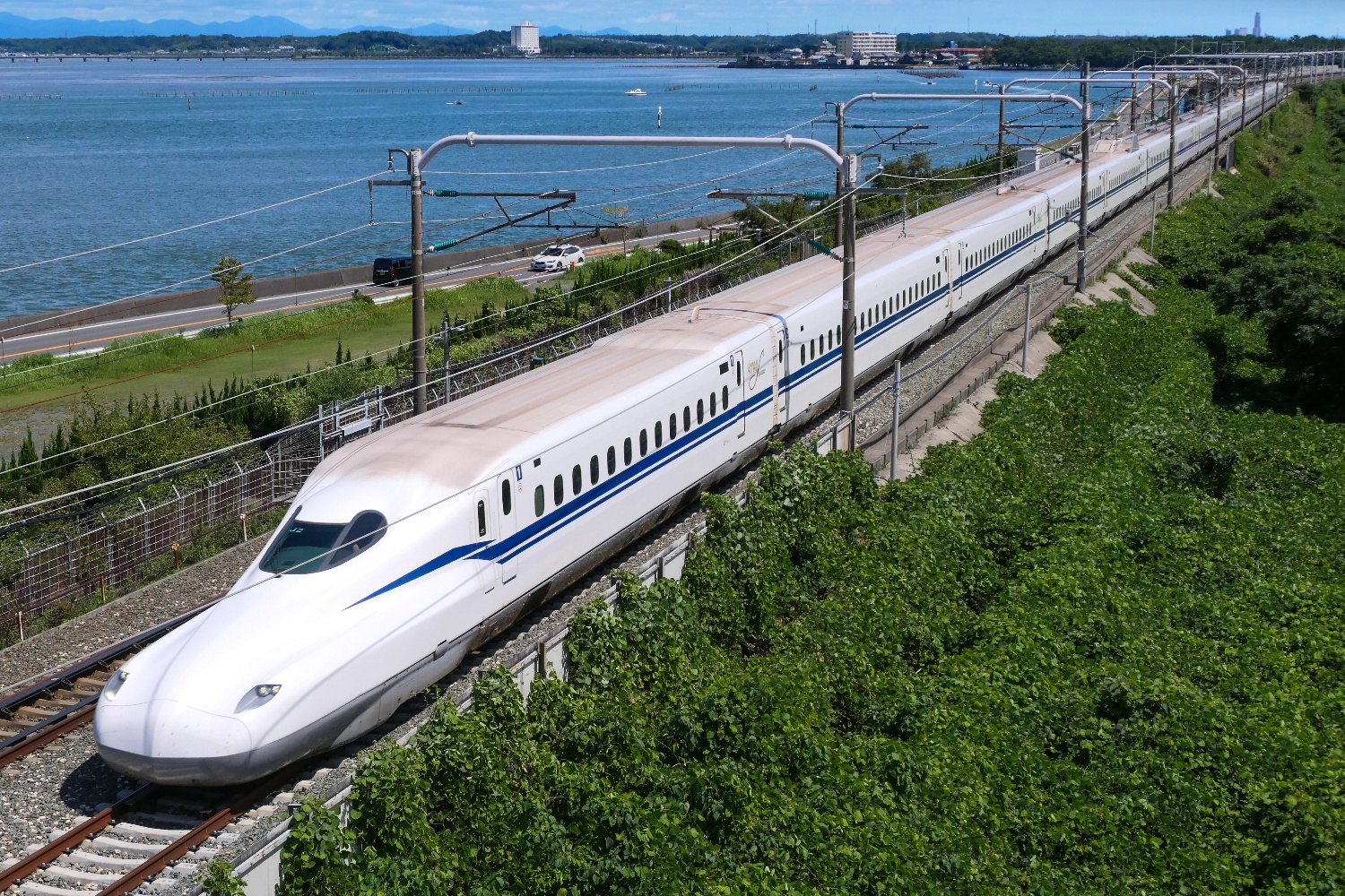 A sleek white Japanese bullet train speeds past lush green foliage beside a vast blue bay.