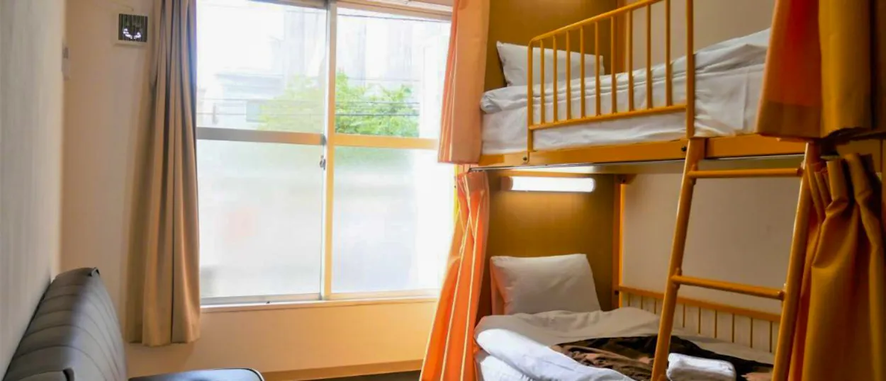 Bright hostel room in Tokyo featuring wooden bunk beds with white bedding and orange privacy curtains.