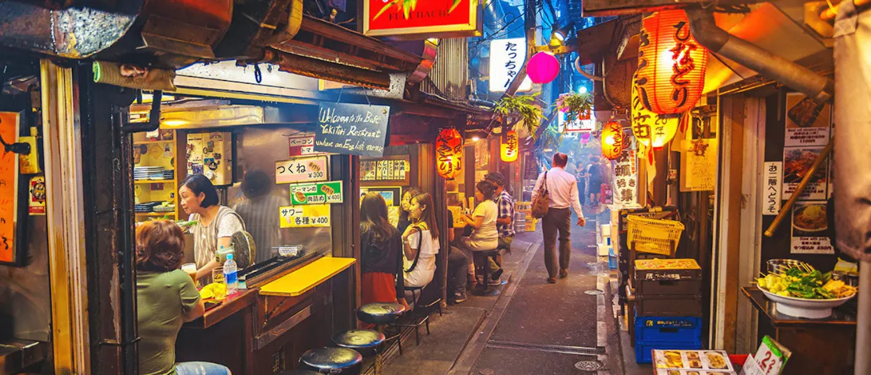 A lively Tokyo alley at night, with people dining at small eateries under glowing lanterns.