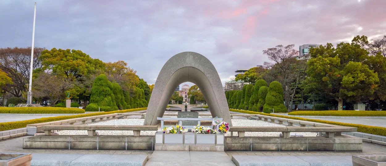 A poignant view of the Hiroshima Peace Memorial Park Cenotaph with flowers, surrounded by green trees.