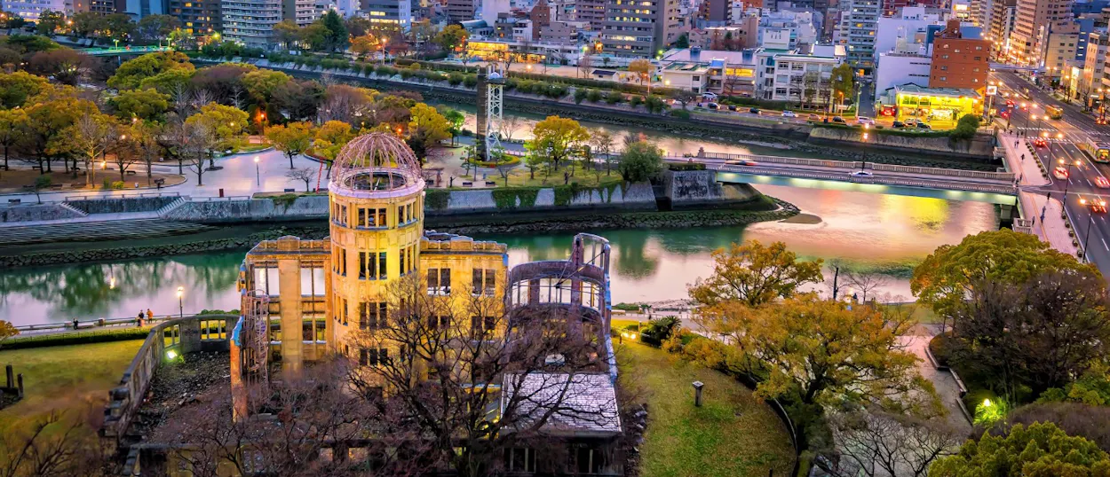 The historic Atomic Bomb Dome stands beside a river in Hiroshima, with city lights illuminating the background.