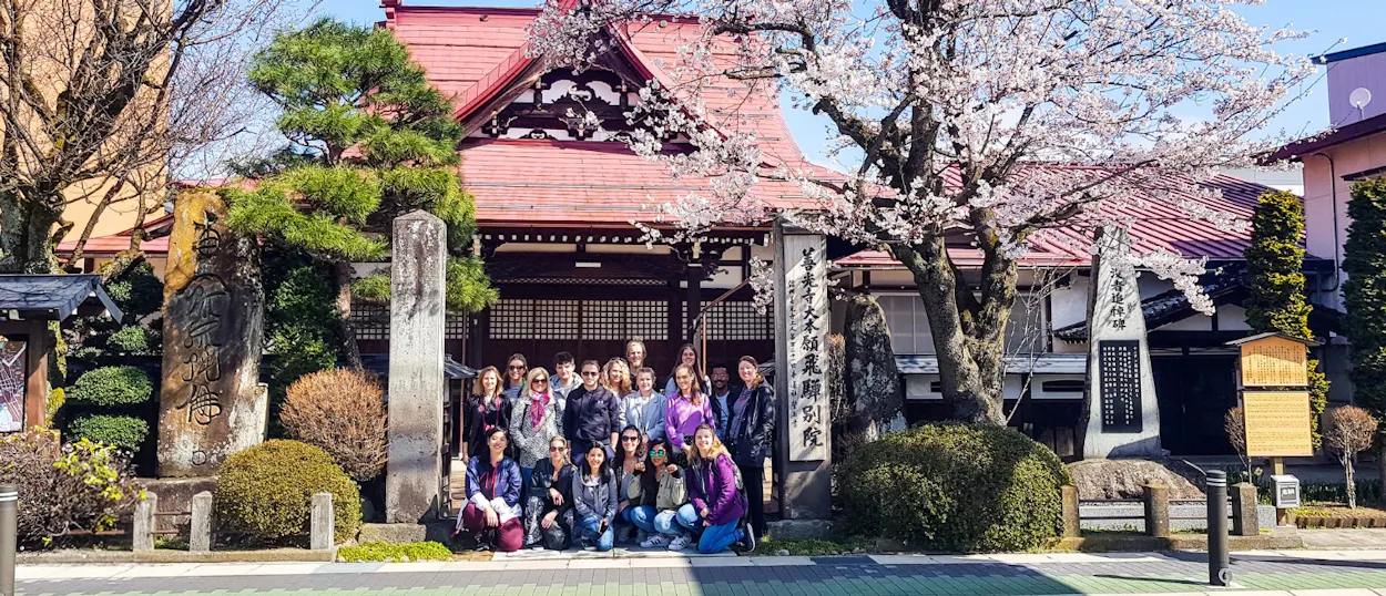 A group of happy backpackers pose in front of a traditional Japanese temple with blooming cherry blossoms.