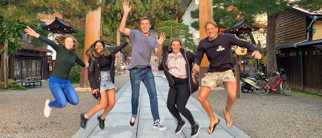 Five joyful backpackers jump high in the air at a traditional Japanese shrine in Tokyo.
