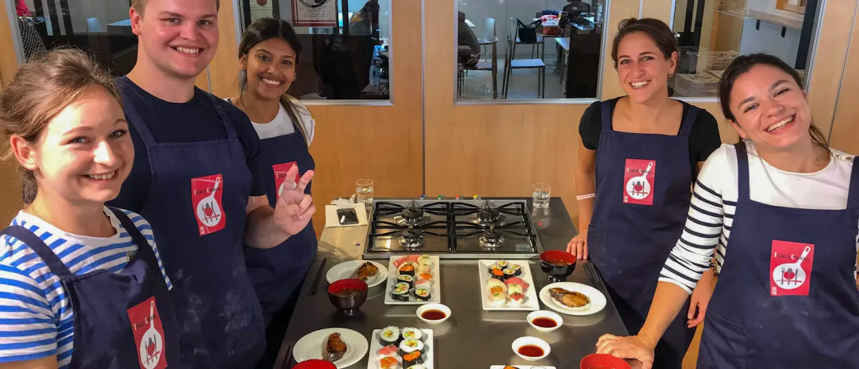 Five smiling travelers in aprons proudly display their handmade sushi and miso soup in Tokyo.