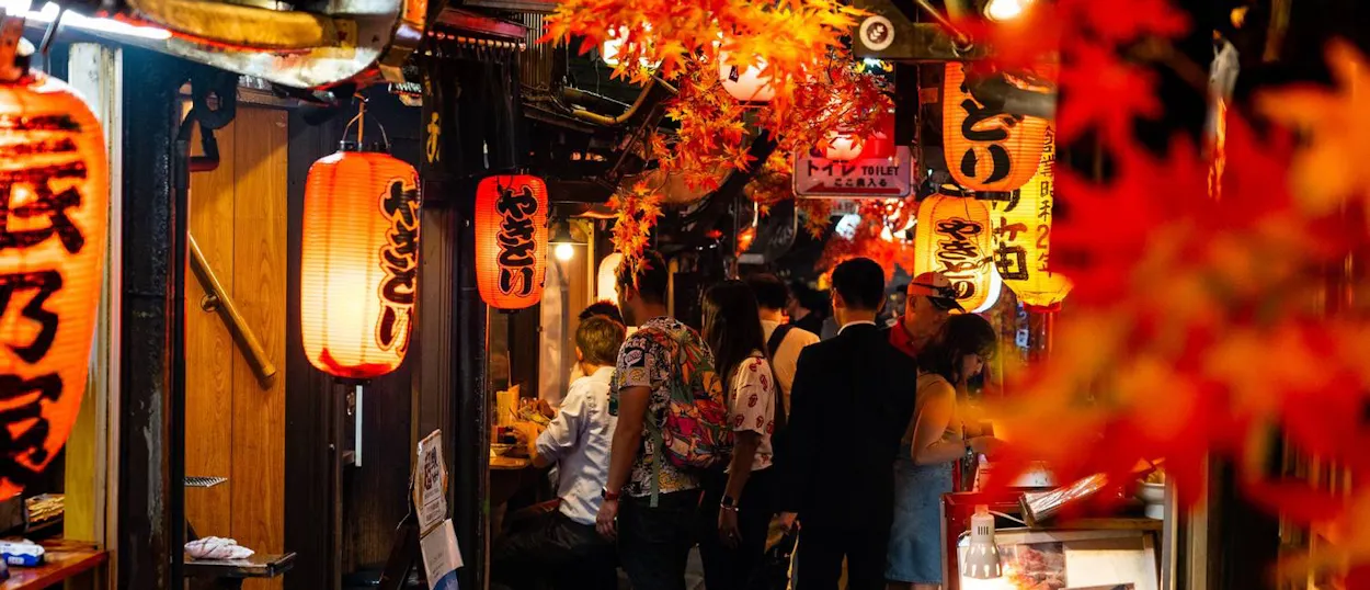 People walk through a vibrant, lantern-lit alley in Shinjuku, Tokyo, adorned with red foliage.