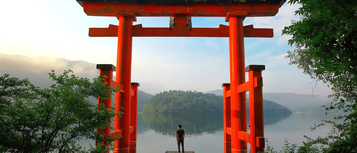 A lone traveler stands beneath a vibrant red torii gate, overlooking a misty lake in Hakone, Japan.