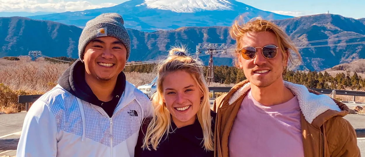 Three smiling friends pose happily outdoors with the majestic, snow-capped Mount Fuji behind them.