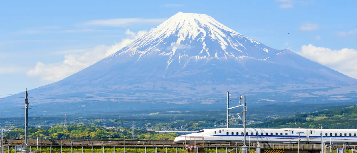 A white Shinkansen bullet train speeds past green trees with majestic, snow-capped Mt. Fuji.