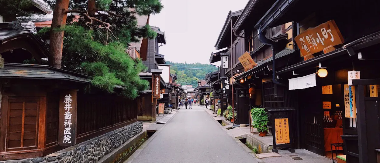 Traditional dark wooden buildings line a quiet street in Takayama, Japan, under an overcast sky.