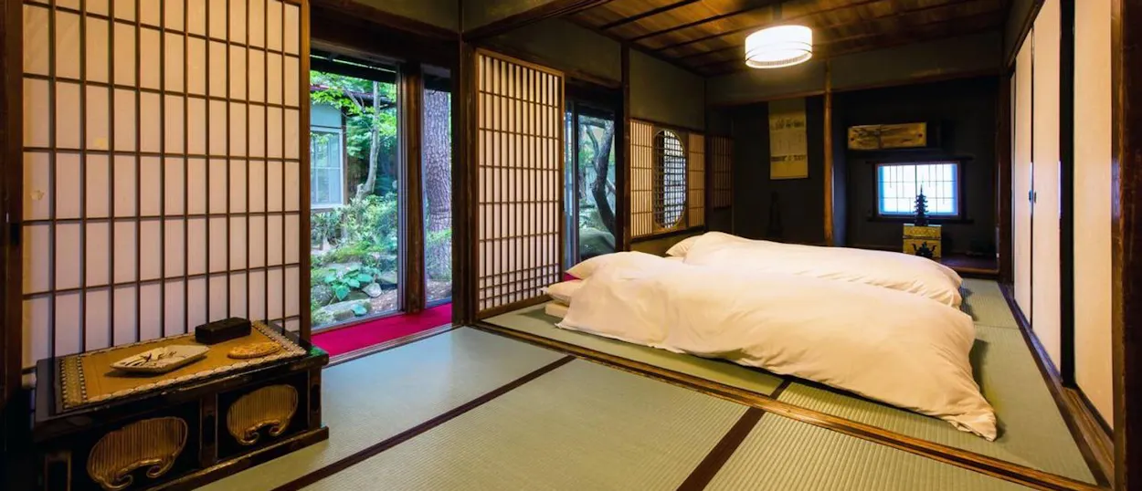A serene Japanese temple room with tatami mats, futon beds, and a view of a lush garden.