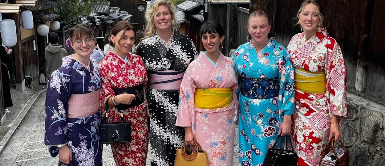 Six tourists smiling in vibrant kimonos on a traditional street in Kyoto's Gion district.