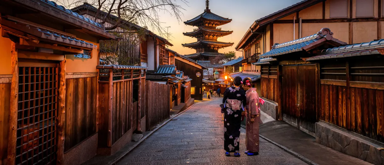 Two women in kimonos stroll a historic Kyoto street towards a beautiful pagoda at dusk.