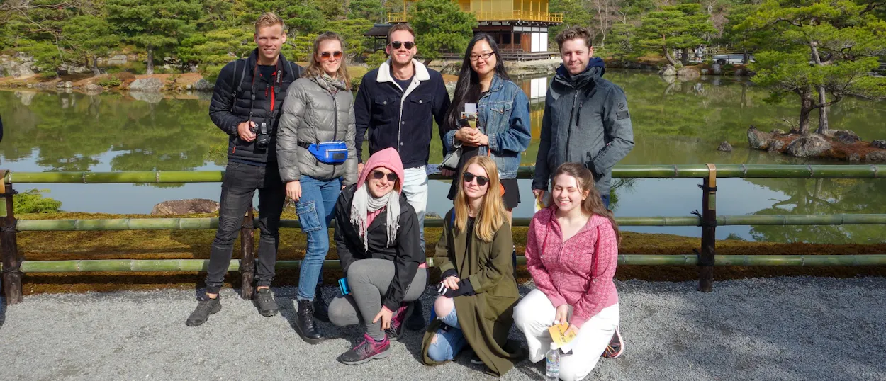 Eight smiling travelers pose by a bamboo fence with Kyoto's golden pavilion and tranquil pond behind them.