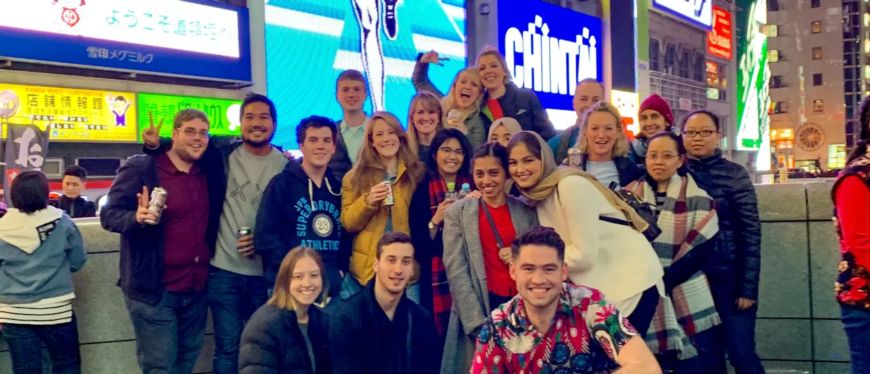 A diverse group of smiling friends enjoys the bright lights and energy of Dotonbori, Osaka.