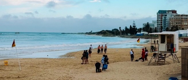 People strolling along a wide sandy beach past a lifeguard tower and safety flags