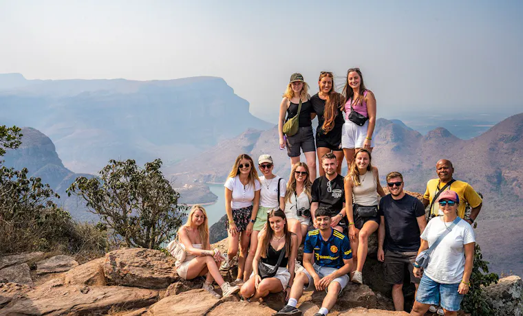 A group of happy backpackers posing on a rocky viewpoint overlooking the vast Blyde River Canyon.