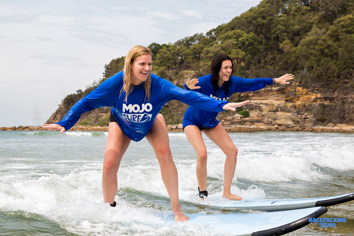 Two smiling women in blue shirts learning to surf on waves at Byron Bay.