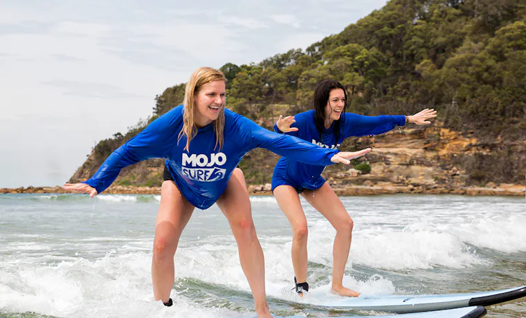 Two smiling women in blue shirts learning to surf on waves at Byron Bay.