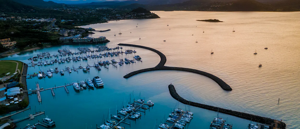 Aerial view of Airlie Beach marina at sunset, with numerous boats and a tranquil coastal town.