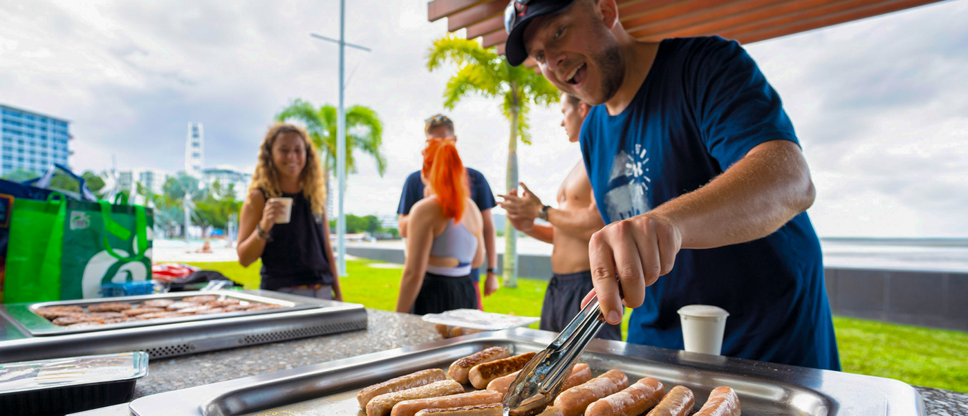 A smiling man grills sausages at an outdoor barbecue in Cairns with friends enjoying the tropical setting.