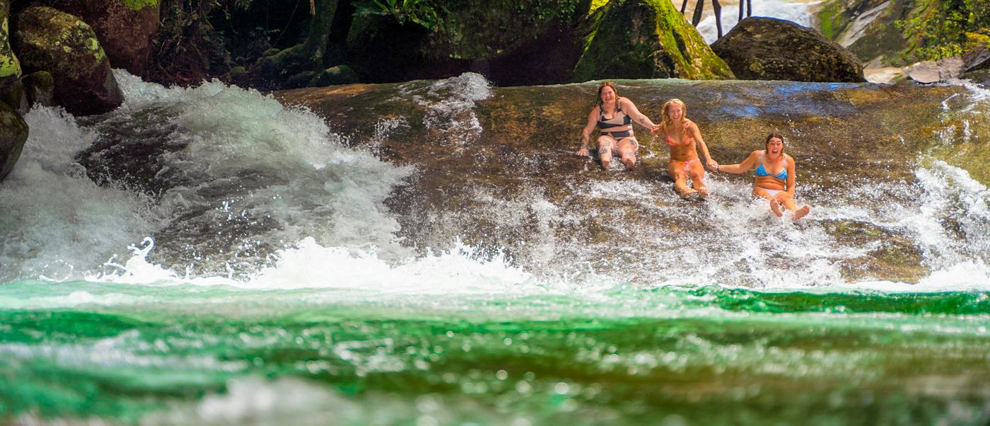 Three women laugh while sliding down a natural rock waterfall into green water at Josephine Falls.