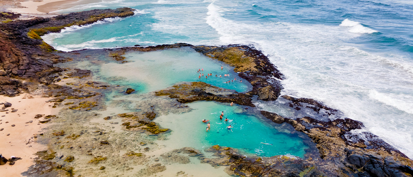 Tourists swim in the natural, clear turquoise Champagne Pools on Fraser Island, surrounded by rocks and ocean.