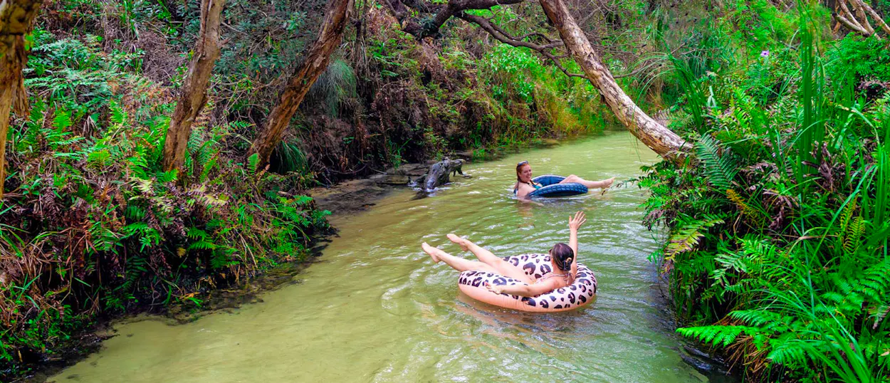Two women happily float on tubes down a lush green creek on K'gari, Australia.