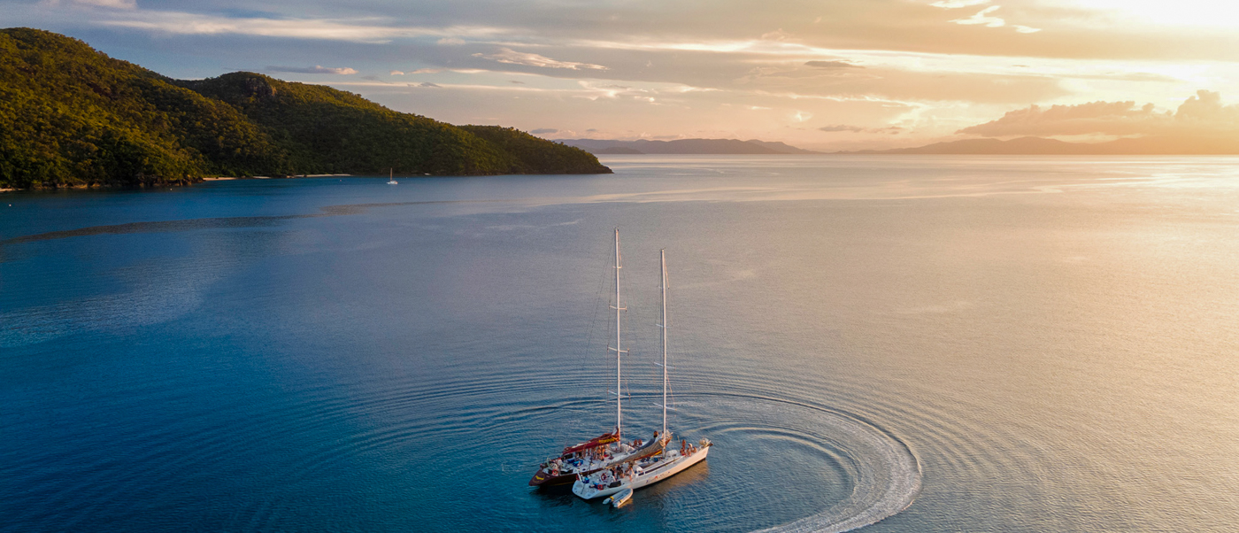 A catamaran sails on calm turquoise water near green islands in the Whitsundays at sunset.