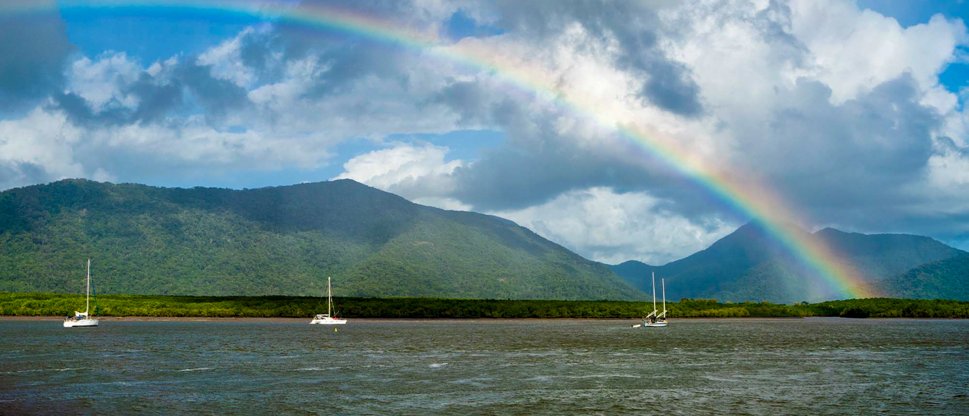 A vibrant rainbow arches over lush tropical mountains and three sailboats in Cairns.