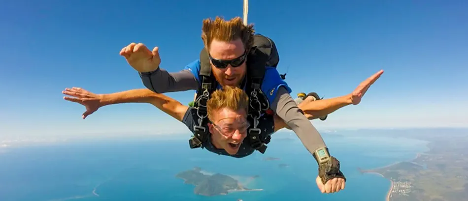 Two men skydive tandem over the blue ocean and coastline near Mission Beach, Australia, with excited expressions.