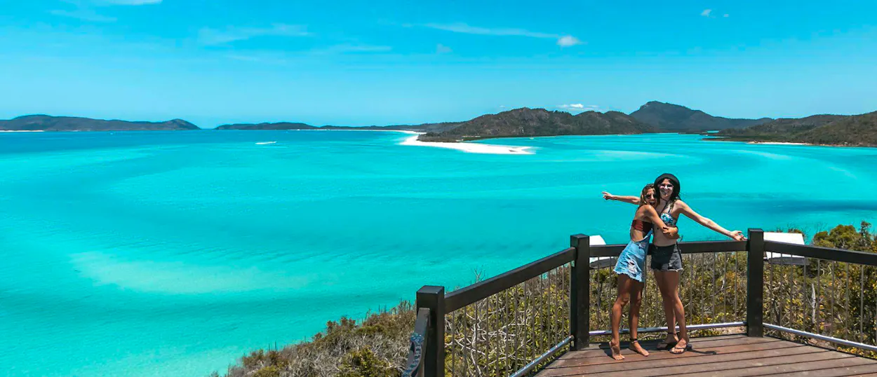 Two friends embrace on a lookout, admiring the panoramic view of Whitehaven Beach in the Whitsundays.