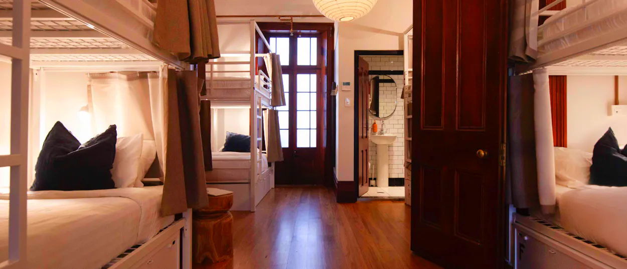 A warmly lit, clean hostel dorm room with white bunk beds and privacy curtains in Sydney.