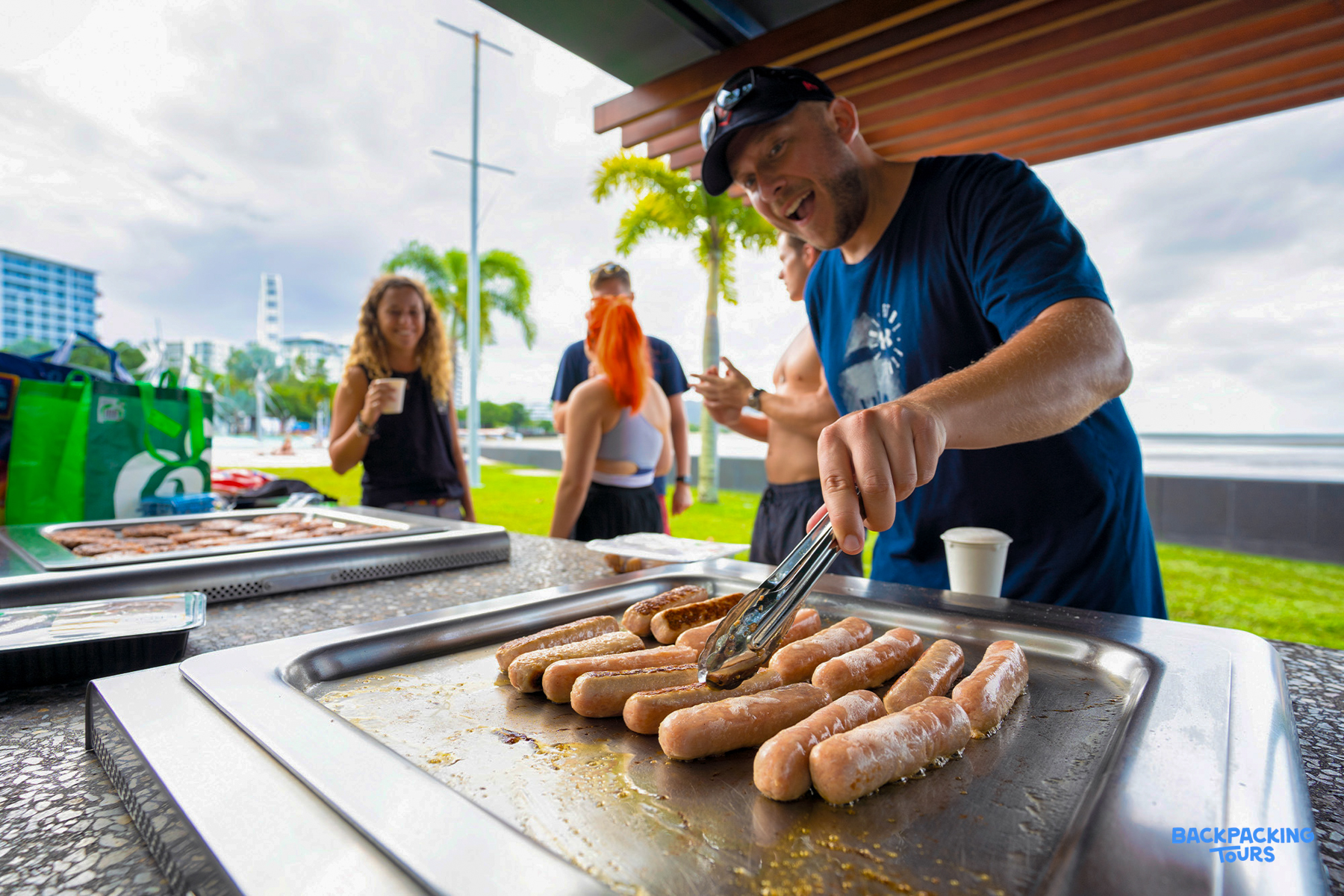 A man happily grills sausages for backpackers at a lively outdoor BBQ in Cairns.