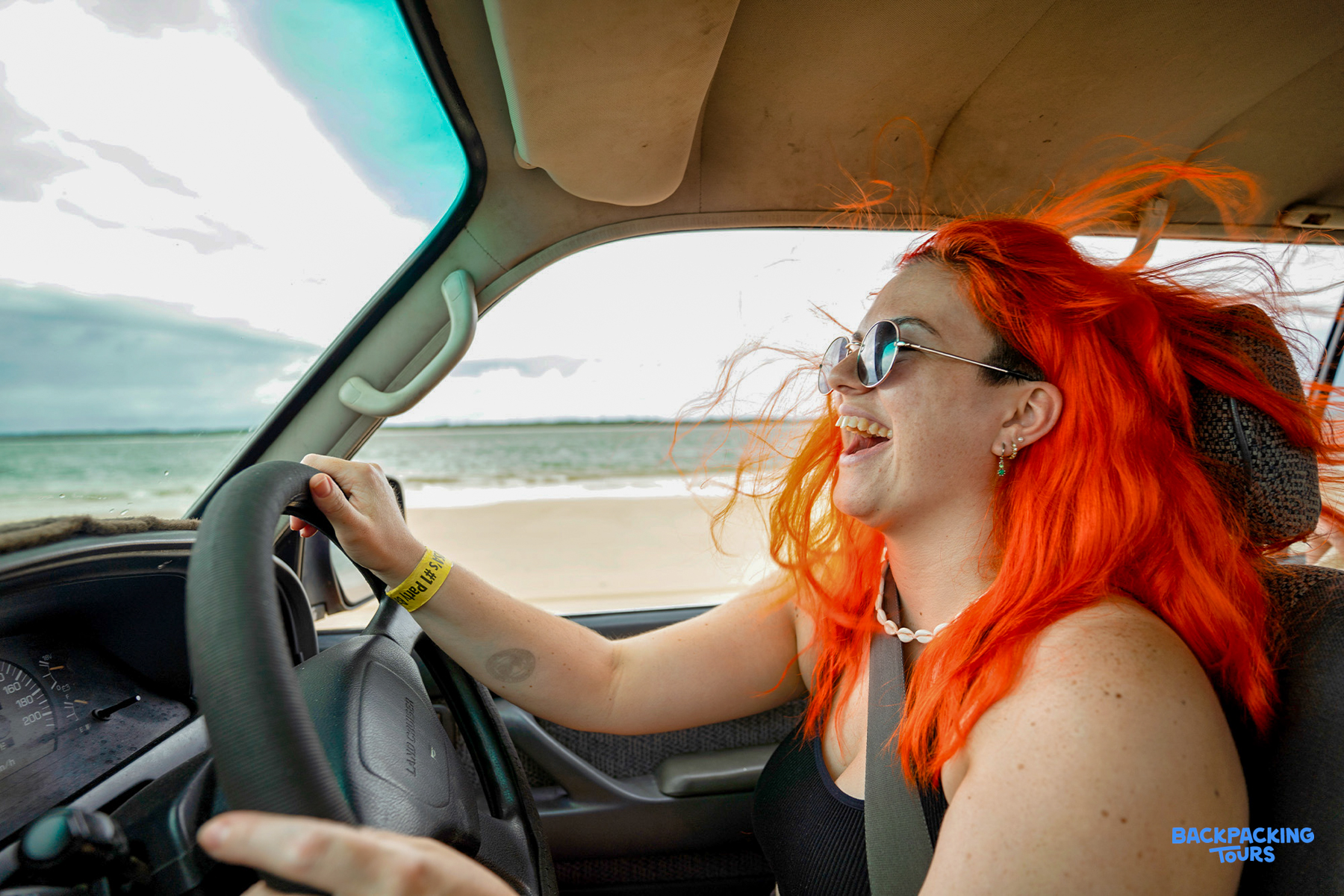 A joyful woman with vibrant orange hair drives a 4WD along Rainbow Beach, smiling.