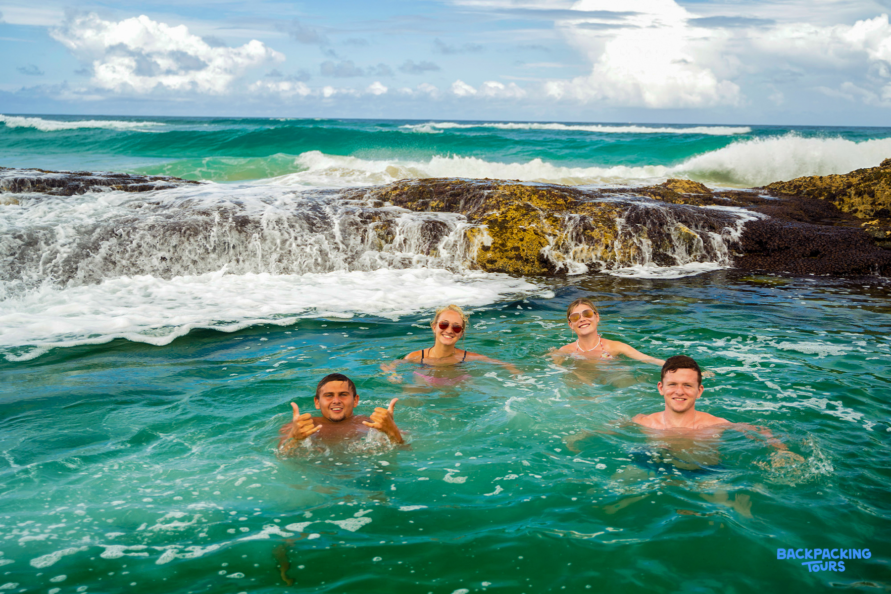 Four happy backpackers swim in the natural Champagne Pools on K'gari, enjoying the ocean waves.