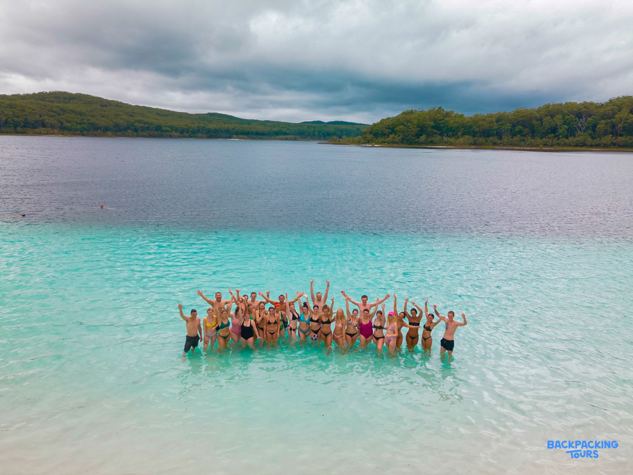 A group of happy backpackers stands in the clear turquoise water of Lake McKenzie, K'gari.