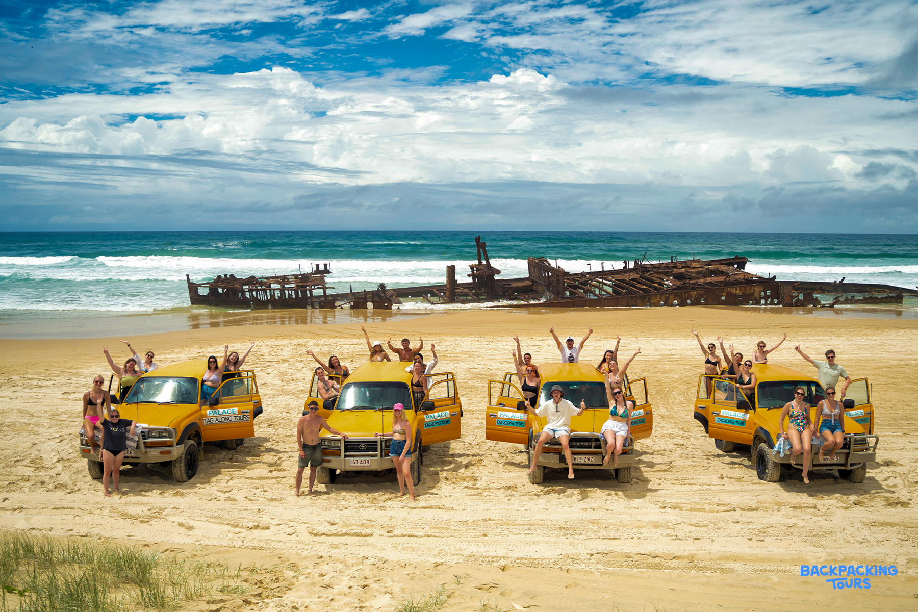 Happy backpackers cheer from 4WDs on Fraser Island beach with the historic Maheno shipwreck.